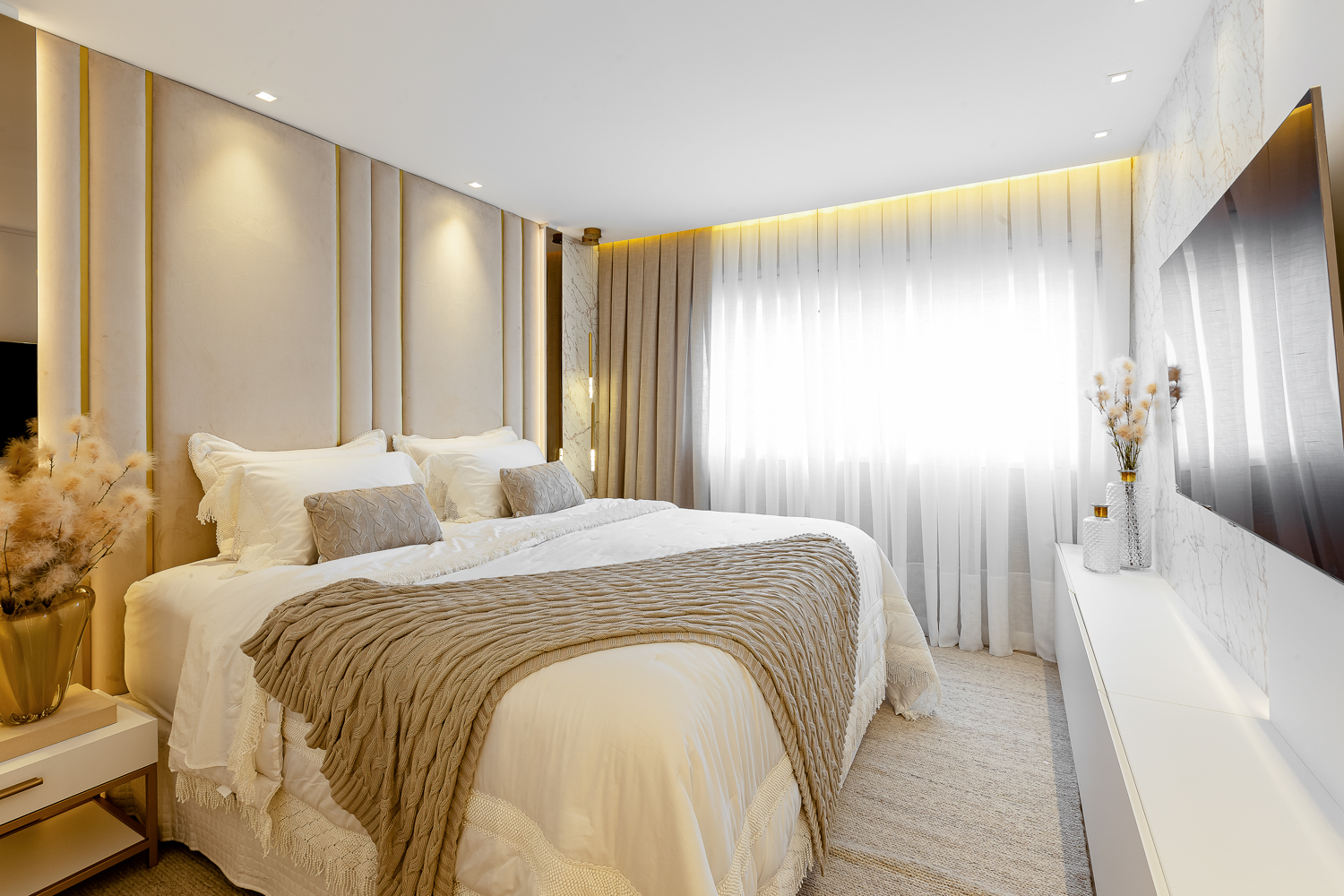 Suíte ampla em tons neutros com cama central e iluminação natural filtrada pelas cortinas.